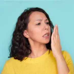 A person with curly hair wears a bright yellow sweater and gestures with their hand against a teal background.