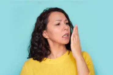 A person with curly hair wears a bright yellow sweater and gestures with their hand against a teal background.