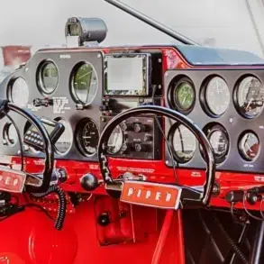 Cockpit view of a Piper plane, featuring black and red controls, gauges, and flight instruments with dual yokes and headsets.