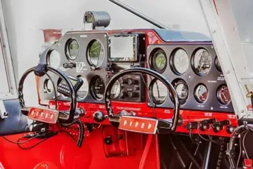 Cockpit view of a Piper plane, featuring black and red controls, gauges, and flight instruments with dual yokes and headsets.