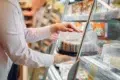 A person in a light-colored shirt is reaching for a cake in a refrigerated display case at a grocery store.