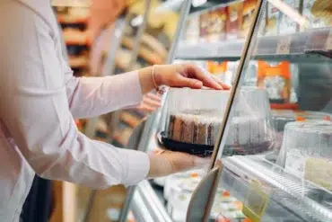 A person in a light-colored shirt is reaching for a cake in a refrigerated display case at a grocery store.