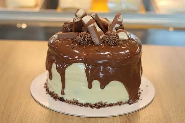 Chocolate-drizzled cake topped with chocolate bars and truffles, on a light wooden surface, showcasing a delicious dessert.