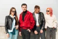 Four stylish young adults stand together, showcasing trendy outfits against a bright, minimalist background.