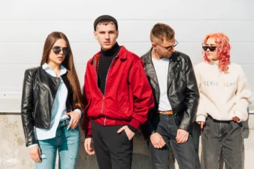 Four stylish young adults stand together, showcasing trendy outfits against a bright, minimalist background.