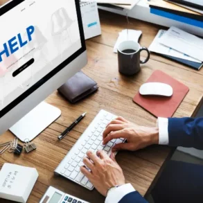 A person's hands typing on a keyboard in front of a desktop computer displaying a "HELP" webpage, with a coffee mug and documents nearby.
