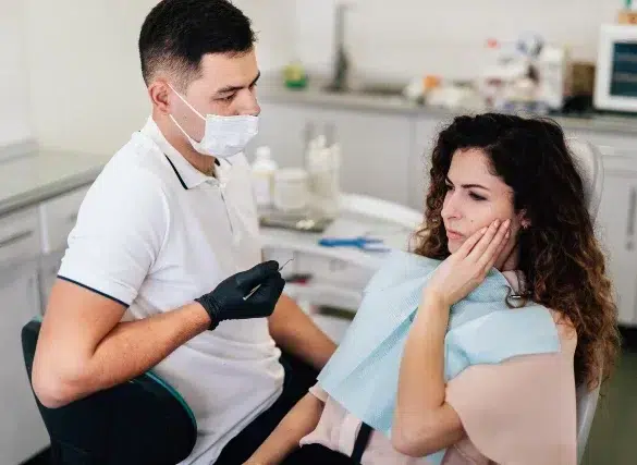 A patient sits in a dental chair, covered with a bib, while a dentist prepares to perform a procedure in a bright clinic.