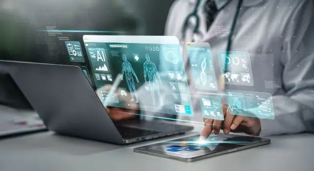 A healthcare professional interacts with holographic medical data displays above a laptop and tablet in a modern workspace.