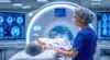 A medical professional in scrubs assists a patient lying on a bed near a CT scan machine, with diagnostic screens in the background.