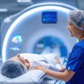 A medical professional in scrubs assists a patient lying on a bed near a CT scan machine, with diagnostic screens in the background.
