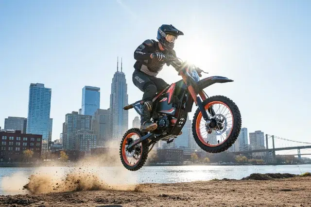 A rider jumps a dirt bike near a riverbank, with a backdrop of a city skyline and dust cloud rising beneath the wheels.