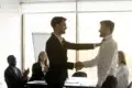Two men are shaking hands in a bright office setting, while colleagues applaud in the background.