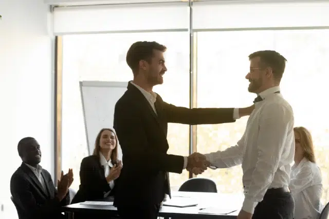 Two men are shaking hands in a bright office setting, while colleagues applaud in the background.