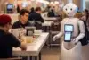 A humanoid robot with a touchscreen tablet stands in a bustling cafeteria, engaging with people seated at tables.