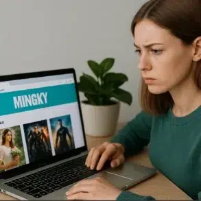 A person in a green sweater uses a laptop displaying a webpage titled "MINGKY" with movie posters on the screen, beside a potted plant.