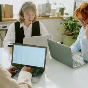 A group of three professionals in headsets is collaborating at a table with laptops in a bright office space.