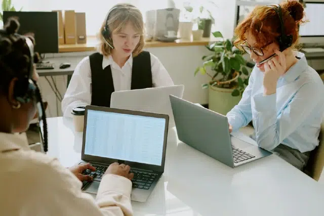 A group of three professionals in headsets is collaborating at a table with laptops in a bright office space.