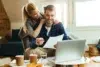 A woman hugs a man while he reviews documents on a laptop at a cozy living room with coffee and snacks on the table.