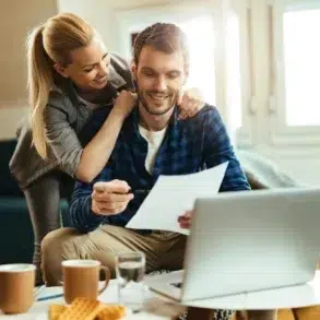 A woman hugs a man while he reviews documents on a laptop at a cozy living room with coffee and snacks on the table.