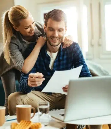A woman hugs a man while he reviews documents on a laptop at a cozy living room with coffee and snacks on the table.