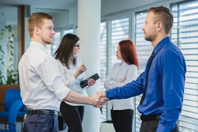 Two men in business attire shake hands in an office setting, while two women observe in the background. Natural light streams through large windows.