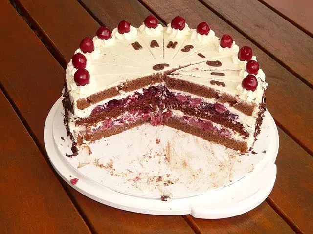 A partially sliced chocolate cake topped with whipped cream and cherries, with "HANS" written on the frosting, on a wooden table.