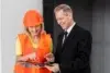 A woman in an orange safety vest and helmet discusses a clipboard with a man in a black suit, highlighting a collaborative work environment.