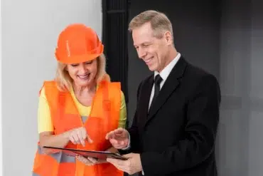A woman in an orange safety vest and helmet discusses a clipboard with a man in a black suit, highlighting a collaborative work environment.