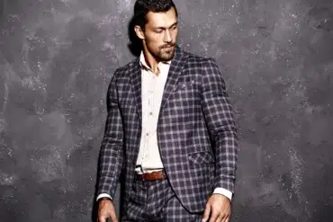 A man in a stylish checked suit stands confidently against a textured gray wall, showcasing modern fashion elegance.