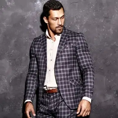 A man in a stylish checked suit stands confidently against a textured gray wall, showcasing modern fashion elegance.