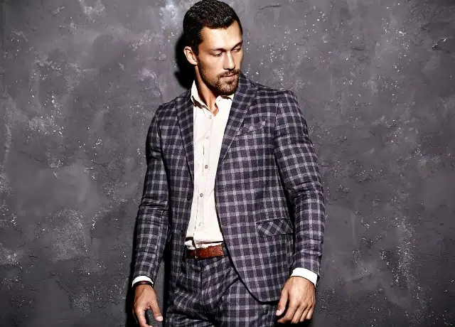A man in a stylish checked suit stands confidently against a textured gray wall, showcasing modern fashion elegance.