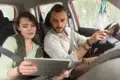 A man is driving a car while a woman beside him holds a tablet, both appearing engaged in their activity.