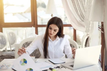 A person in a white shirt reviews charts and data on a table with a laptop, in a bright, stylish café setting.