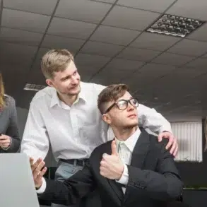 A group of five office workers collaborates around a laptop, with one person giving a thumbs-up in a modern workspace.