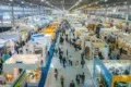 Aerial view of a busy trade exhibition hall filled with colorful booths and crowds of attendees exploring various displays.