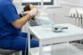A dentist in blue scrubs performs a procedure on a patient in a dental office, with dental equipment and tools visible nearby.