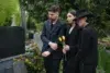 Three individuals dressed in black pay their respects in a cemetery, one holding yellow tulips, surrounded by greenery and gravestones.