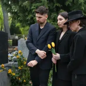 Three individuals dressed in black pay their respects in a cemetery, one holding yellow tulips, surrounded by greenery and gravestones.