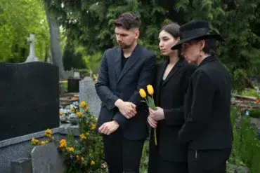 Three individuals dressed in black pay their respects in a cemetery, one holding yellow tulips, surrounded by greenery and gravestones.