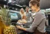 A cashier processes a payment at a register, with a pineapple visible on the counter and customers nearby. Brightly lit store interior.