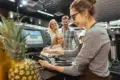 A cashier processes a payment at a register, with a pineapple visible on the counter and customers nearby. Brightly lit store interior.