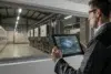 A worker monitors industrial machinery through a window, using a tablet to display real-time data and analytics.