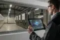 A worker monitors industrial machinery through a window, using a tablet to display real-time data and analytics.