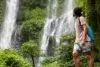 A man stands in awe before a lush waterfall, surrounded by greenery, with a backpack slung over his shoulder.