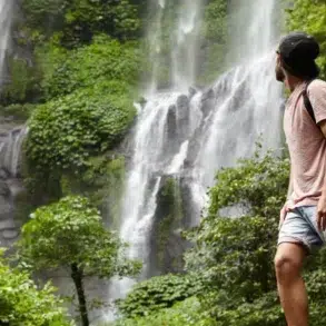 A man stands in awe before a lush waterfall, surrounded by greenery, with a backpack slung over his shoulder.