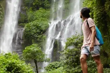 A man stands in awe before a lush waterfall, surrounded by greenery, with a backpack slung over his shoulder.