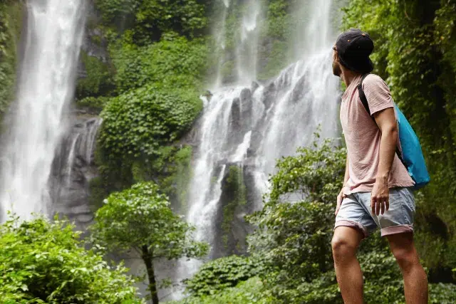 A man stands in awe before a lush waterfall, surrounded by greenery, with a backpack slung over his shoulder.