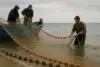 Three fishermen pulling a fishing net from the water, with a blue boat anchored nearby, under a cloudy sky.