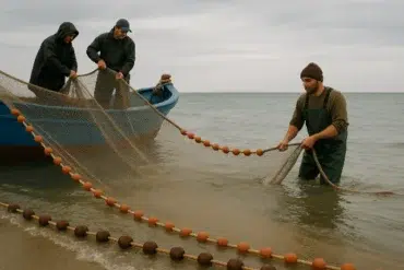 Three fishermen pulling a fishing net from the water, with a blue boat anchored nearby, under a cloudy sky.
