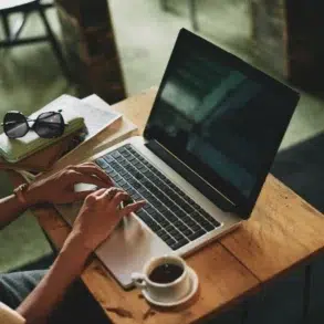 A person typing on a laptop at a wooden table, surrounded by books, sunglasses, and a cup of coffee in a cozy café setting.